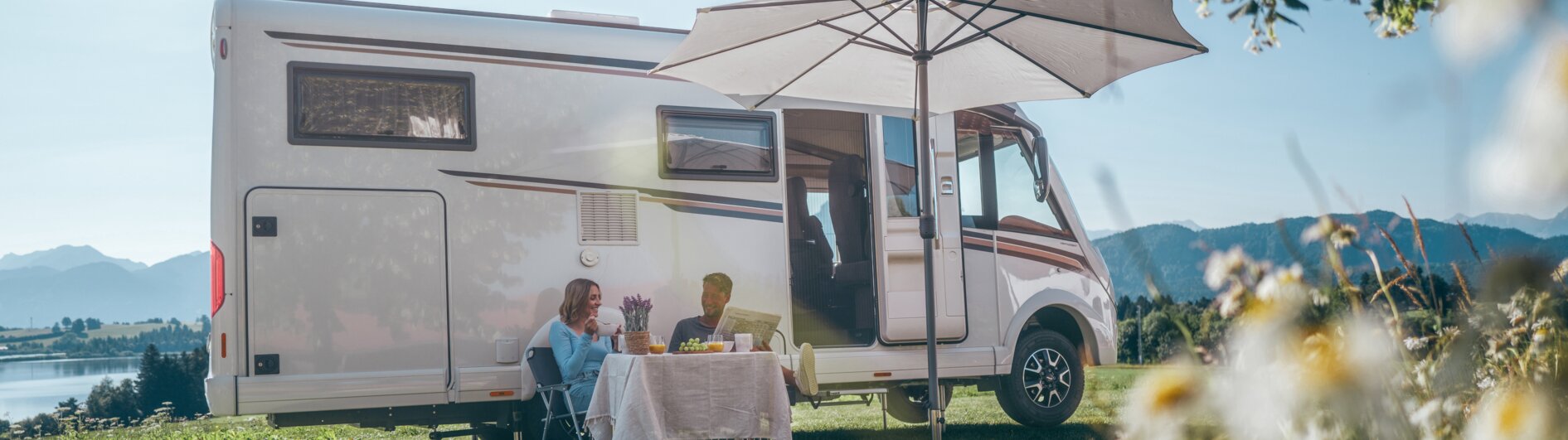Ein Mann und eine Frau sitzen vor ihrem Campervan unter dem Sonnenschirm und trinken Kaffee.
